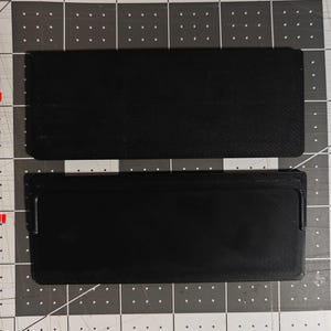 May include: Two black rectangular objects, one labeled "BACK" and the other "FRONT", are displayed on a gray and black grid surface. The objects appear to be made of a textured material, possibly fabric or a similar material.