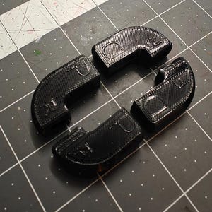 May include: Four black, curved, plastic components with a textured surface are arranged on a black and gray grid. Each piece has a distinct shape with rounded corners and a small, circular detail. The items appear to be 3D-printed.