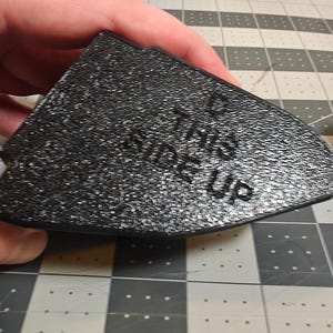 May include: A black, textured, wedge-shaped object with the words THIS SIDE UP embossed on the surface. The object has a flat top and a curved front edge, with a secondary black component visible to the right side of the main piece.