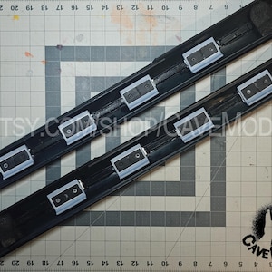 May include: Two black automotive trim pieces with multiple silver and black rectangular components. The pieces are positioned on a gray cutting mat with measurements. The text "ETSY.COM/SHOP/CAVEMODS" is visible. The Cave Mods logo is in the lower right corner.