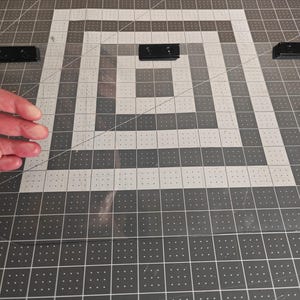 May include: A clear, rectangular acrylic sheet with three black mounting brackets. The sheet is positioned on a gray and white grid surface. The image shows a hand near the sheet.