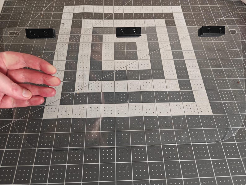 May include: A clear, rectangular acrylic sheet with three black mounting brackets. The sheet is positioned on a gray and white grid surface. The image shows a hand near the sheet.