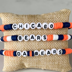 May include: Three beaded bracelets with the words "CHICAGO", "BEARS", and "DA BEARS" spelled out in white letter beads. The bracelets feature orange, navy blue, and white beads. They are displayed on a burlap pillow.
