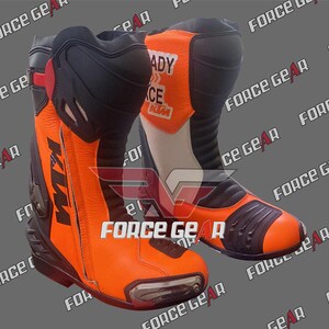 Könnte beinhalten: Paar orange-schwarze Motorradstiefel mit KTM-Logo. Die Stiefel haben einen Reißverschluss, eine verstärkte Spitze und Schutzpaneele. Der Text "READY RACE KTM" ist auf dem oberen Schaft sichtbar. Die Stiefel sind für den Rennsport konzipiert.