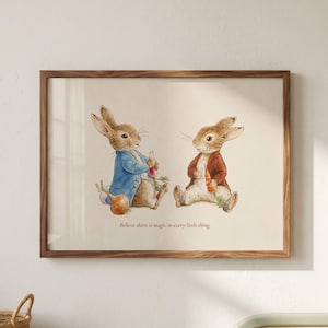 May include: A framed watercolor print featuring two rabbits in clothing, one in a blue jacket and one in a red jacket, holding vegetables. Below the rabbits, the text reads, "Believe there is magic in every little thing." The frame is wood.