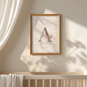 May include: A rectangular wall art print in a gold frame featuring a large dark brown letter A decorated with a soft pink bow. Below the letter, the name AMELIA is printed in dark brown text. The background has a faint floral pattern.