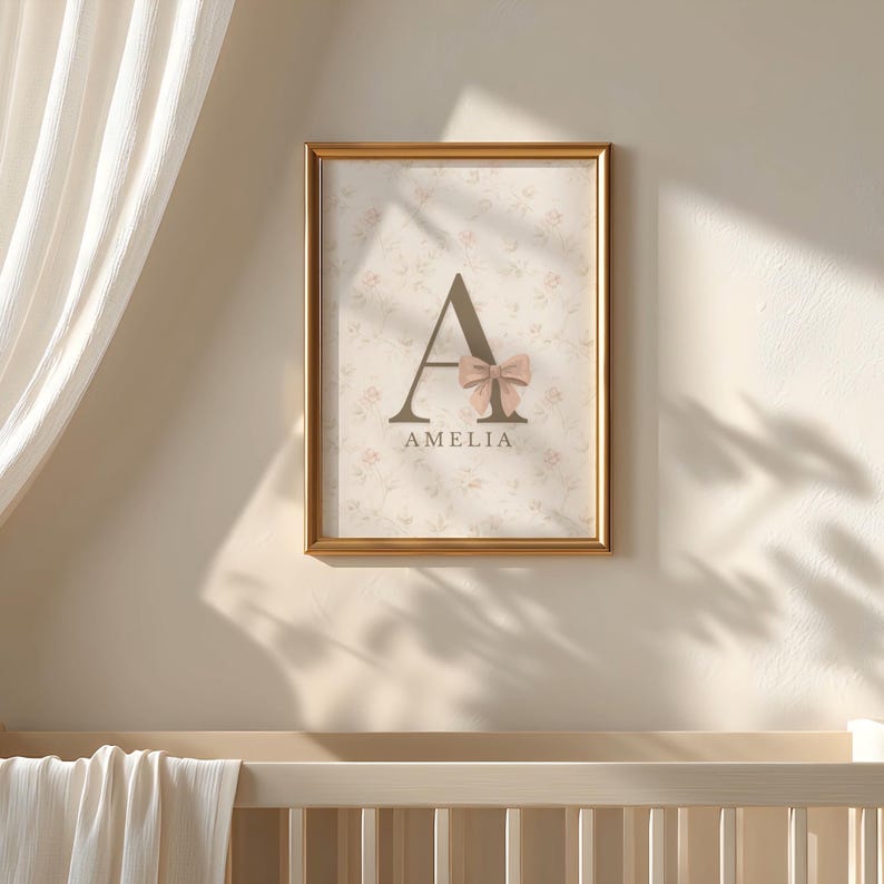 May include: A rectangular wall art print in a gold frame featuring a large dark brown letter A decorated with a soft pink bow. Below the letter, the name AMELIA is printed in dark brown text. The background has a faint floral pattern.