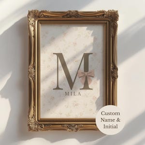 May include: A rectangular wall art print in an ornate gold frame. The print features a large dark brown letter M with a small beige bow attached, the name Mila printed below, and a light cream background with a faint floral pattern.
