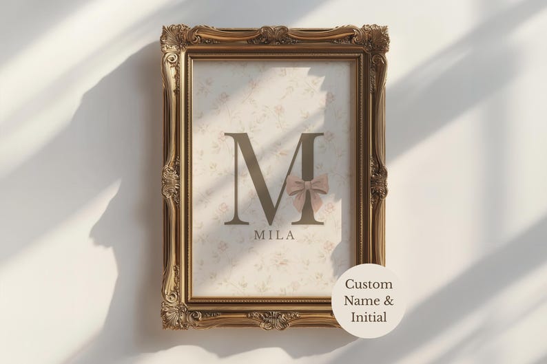 May include: A rectangular wall art print in an ornate gold frame. The print features a large dark brown letter M with a small beige bow attached, the name Mila printed below, and a light cream background with a faint floral pattern.