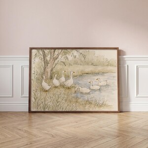 May include: A framed watercolor print depicting a group of white geese near a pond. Three geese stand on the grassy bank near a tree, while three others swim in the water. The scene is rendered in soft, muted earth tones and light green hues.