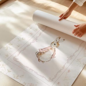 May include: A rolled-up white poster featuring a watercolor illustration of a brown and white rabbit wearing a pink jacket. The rabbit is centered within a decorative rectangular frame, surrounded by a subtle floral pattern on a white background.