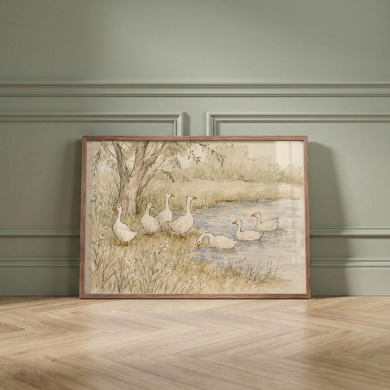 May include: A framed watercolor print featuring a group of white geese near a pond. The scene depicts several geese standing on the grassy bank near a tree and others swimming in the water, rendered in soft muted tones of green, brown, and blue.