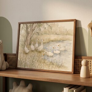 May include: A framed watercolor print depicting a group of white geese near a pond. The scene features several geese standing on the grassy bank under a tree and others swimming in the water, rendered in soft muted tones of green, brown, and blue.