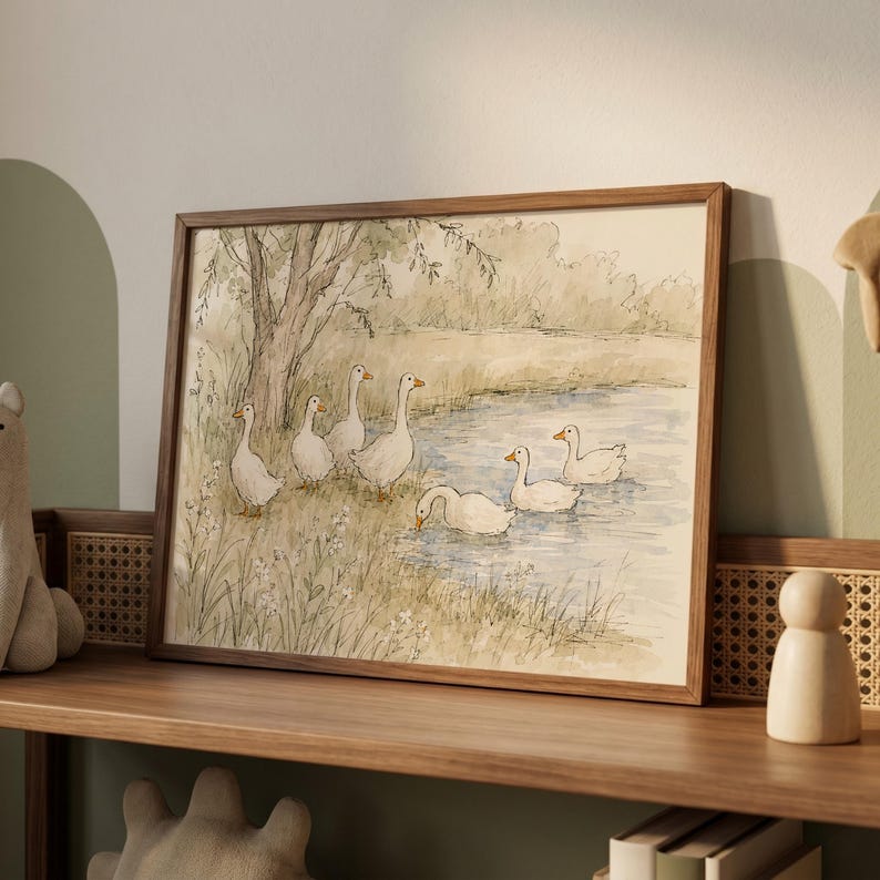 May include: A framed watercolor print depicting a group of white geese near a pond. The scene features several geese standing on the grassy bank under a tree and others swimming in the water, rendered in soft muted tones of green, brown, and blue.