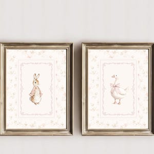 May include: Two framed wall art prints featuring watercolor illustrations. The left print shows a brown rabbit wearing a pink jacket. The right print shows a white goose with a pink bow. Both prints have a light pink floral border and ornate frames.