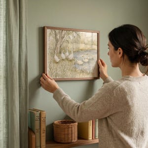 May include: A rectangular wall art print in a dark wood frame featuring a watercolor illustration of a group of white geese near a pond and trees. The artwork depicts several geese standing on a grassy bank and swimming in the water.