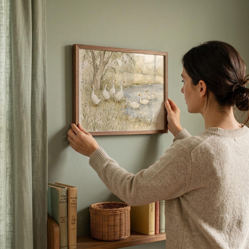 May include: A rectangular wall art print in a dark wood frame featuring a watercolor illustration of a group of white geese near a pond and trees. The artwork depicts several geese standing on a grassy bank and swimming in the water.