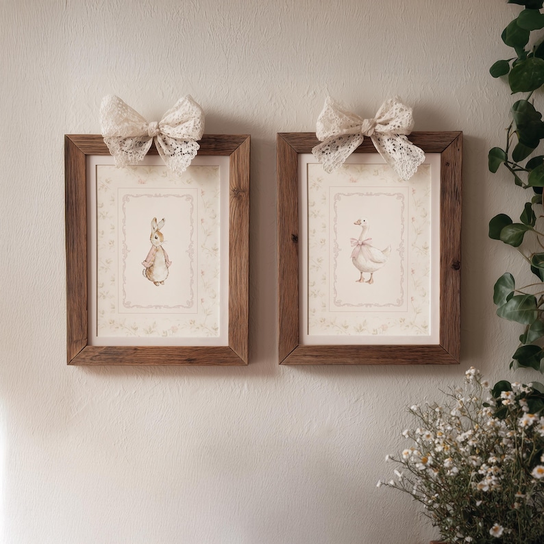 May include: Two rectangular wooden frames containing prints of a rabbit and a goose, each wearing a pink bow. Each frame is topped with a decorative white lace bow. The prints feature a light cream background with a subtle floral border design.