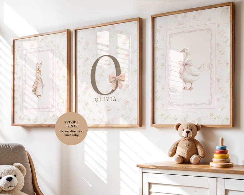 May include: A set of three framed wall prints featuring a rabbit, the letter O with the name OLIVIA and a pink bow, and a goose with a pink bow. Each print has a floral background pattern and is enclosed in a light wood frame for wall display.