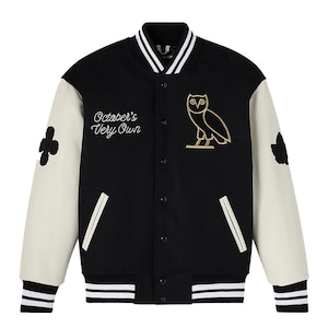 Könnte beinhalten: Schwarze Collegejacke mit cremefarbenen Ärmeln. Mit dem Text "October's Very Own" und einem goldenen Eulenemblem. Enthält schwarz-weiß gestreifte Bündchen und Kragen. Ein Kleeblatt-Patch befindet sich auf dem linken Ärmel.