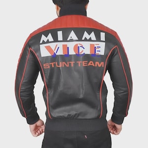 Puede incluir: Chaqueta de cuero negra con detalles rojos en hombros y mangas. La espalda muestra el texto "MIAMI VICE STUNT TEAM" en blanco, naranja y azul. Cuello y puños acanalados.
