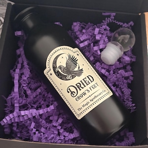 May include: A black bottle labelled "Dried Crow's Feet" with a raven illustration, presented in a box with purple shredded paper. The bottle has a clear glass stopper and is from "The Magic Apothecary Co."