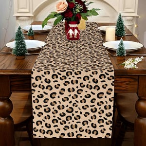 May include: A rectangular table runner featuring a repeating leopard print pattern in shades of black, dark brown, and tan. The fabric runner is laid flat across the center of a wooden dining table, extending from the front edge toward the back.