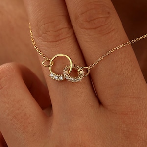 May include: A delicate gold necklace featuring two interlocking circles. One circle is adorned with small, sparkling stones. The necklace is displayed on a person's hand, showcasing its fine details and elegant design.