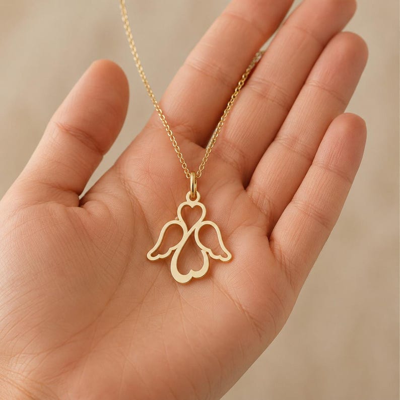 May include: A gold-toned necklace featuring a pendant shaped like a stylised angel with wings and a heart-shaped body. The pendant is suspended from a delicate gold-toned chain, showcasing a cut-out design that forms the silhouette of an angel.