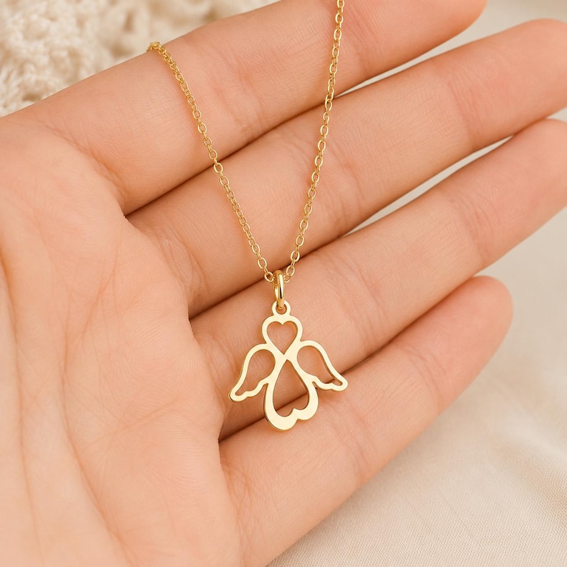 May include: A gold-toned necklace featuring a pendant shaped like an angel with wings and a heart-shaped body. The pendant is suspended from a delicate, thin gold-toned chain. The design is open-work, creating a silhouette of an angel figure.