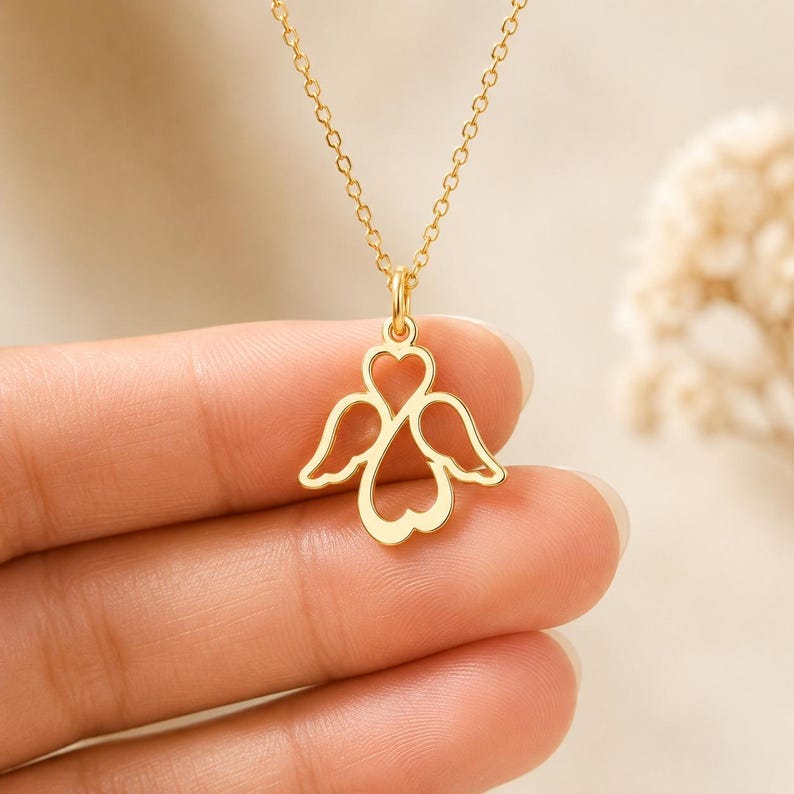 May include: A gold-toned necklace featuring a pendant shaped like a heart with stylised angel wings extending from the sides. The pendant is suspended from a delicate gold-toned chain, showcasing a polished finish and a symmetrical design.