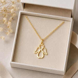 May include: A gold-toned necklace featuring a pendant shaped like an angel with wings and a heart-shaped body. The delicate chain is attached to the top of the pendant, which is displayed inside a white gift box with a light-coloured interior lining.