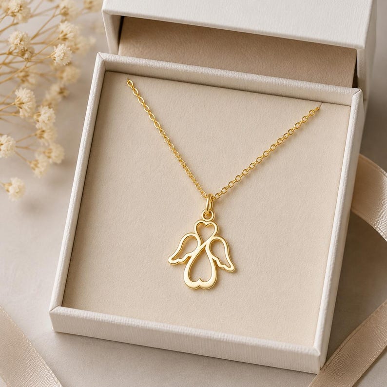 May include: A gold-toned necklace featuring a pendant shaped like an angel with wings and a heart-shaped body. The delicate chain is attached to the top of the pendant, which is displayed inside a white gift box with a light-coloured interior lining.