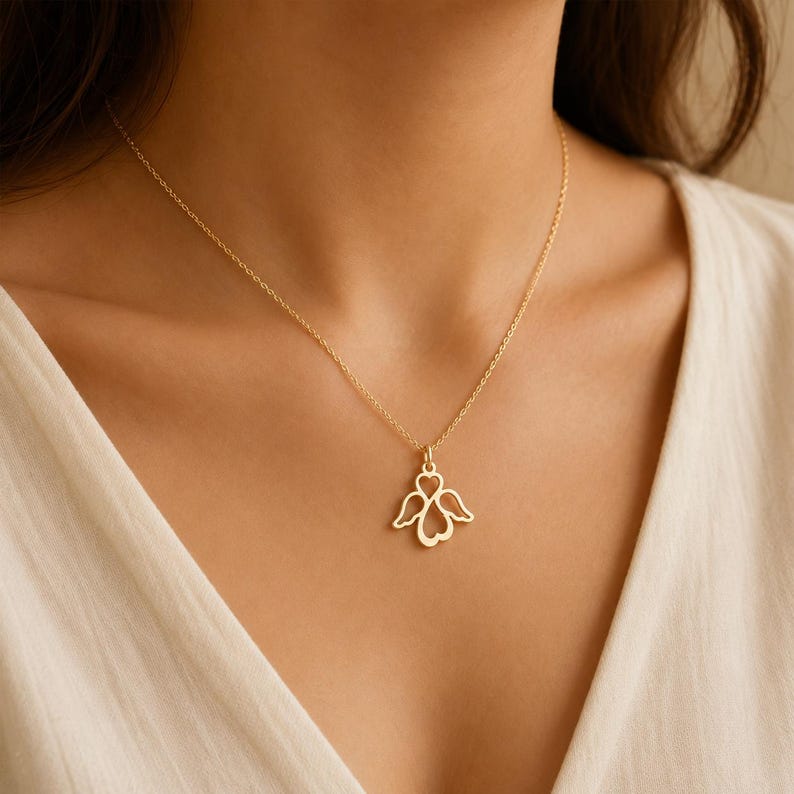 May include: A gold-toned necklace featuring a delicate chain and a pendant shaped like a stylised heart with two symmetrical wings extending from the sides. The pendant is crafted with a thin, open-work metal design, suspended from a simple link chain.