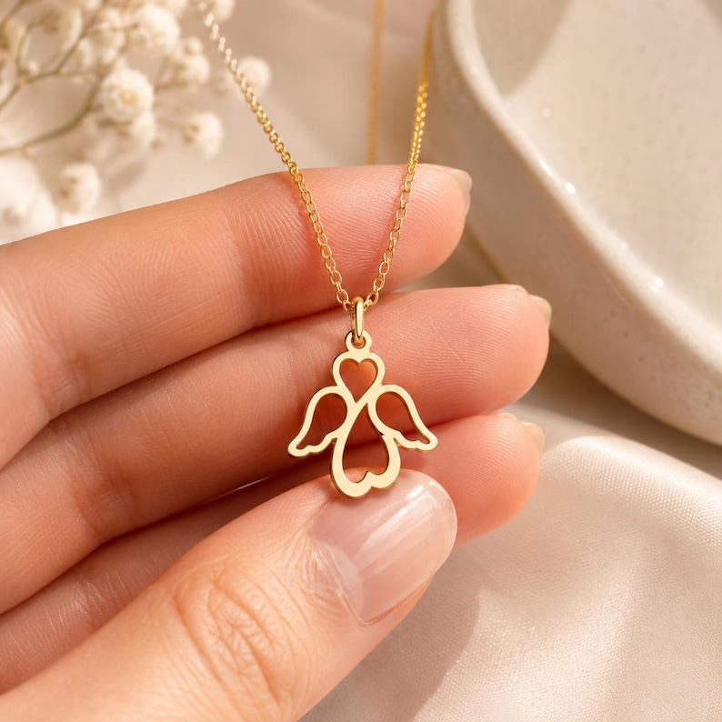 May include: A gold-toned necklace featuring a delicate pendant shaped like an angel with wings formed by open heart outlines. The pendant hangs from a thin, matching gold-toned chain, showcasing a minimalist design with clean, symmetrical lines.