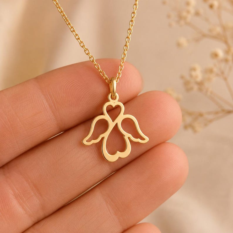 May include: A gold-coloured necklace featuring a pendant shaped like an angel with wings and a heart-shaped centre. The pendant is suspended from a delicate gold-coloured chain. The design is symmetrical, with open spaces forming the wings and heart.