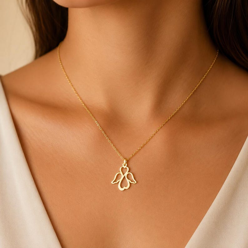 May include: A gold-toned necklace featuring a delicate pendant shaped like an angel with open wings and a heart-shaped body. The pendant is suspended from a thin, matching gold-toned chain that rests against the collarbone area.