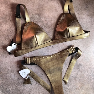 May include: A gold-colored lingerie set featuring a triangle-shaped bra and a thong-style bottom. The bra has adjustable straps, and the thong has a belt-like design with studded details and a buckle closure. Both pieces are made of a shiny, textured material.