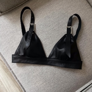 May include: A black leather bralette with adjustable straps and silver-colored buckles. The bralette has a triangular shape with a wide band at the bottom. The straps are attached to the cups with buckles.