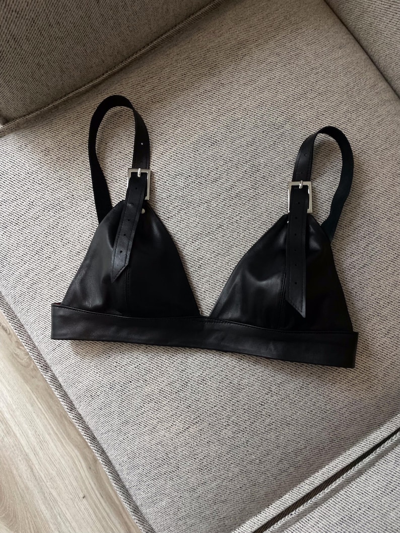 May include: A black leather bralette with adjustable straps and silver-colored buckles. The bralette has a triangular shape with a wide band at the bottom. The straps are attached to the cups with buckles.
