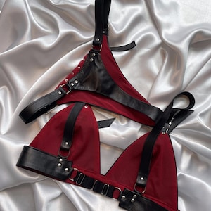 May include: A red and black lingerie set. The set includes a triangle-style bra and a thong, both featuring black leather straps and accents. The fabric is a deep red, and the leather is black. The set is displayed on a white satin surface.