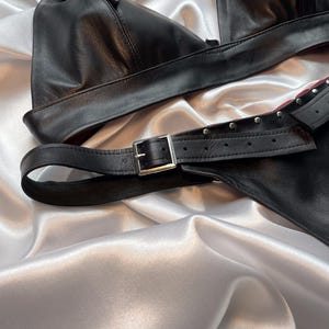 May include: Black leather lingerie set with adjustable straps and silver-tone buckles. The set includes a triangle-shaped top and a matching bottom piece. The lingerie is displayed on a white satin surface.