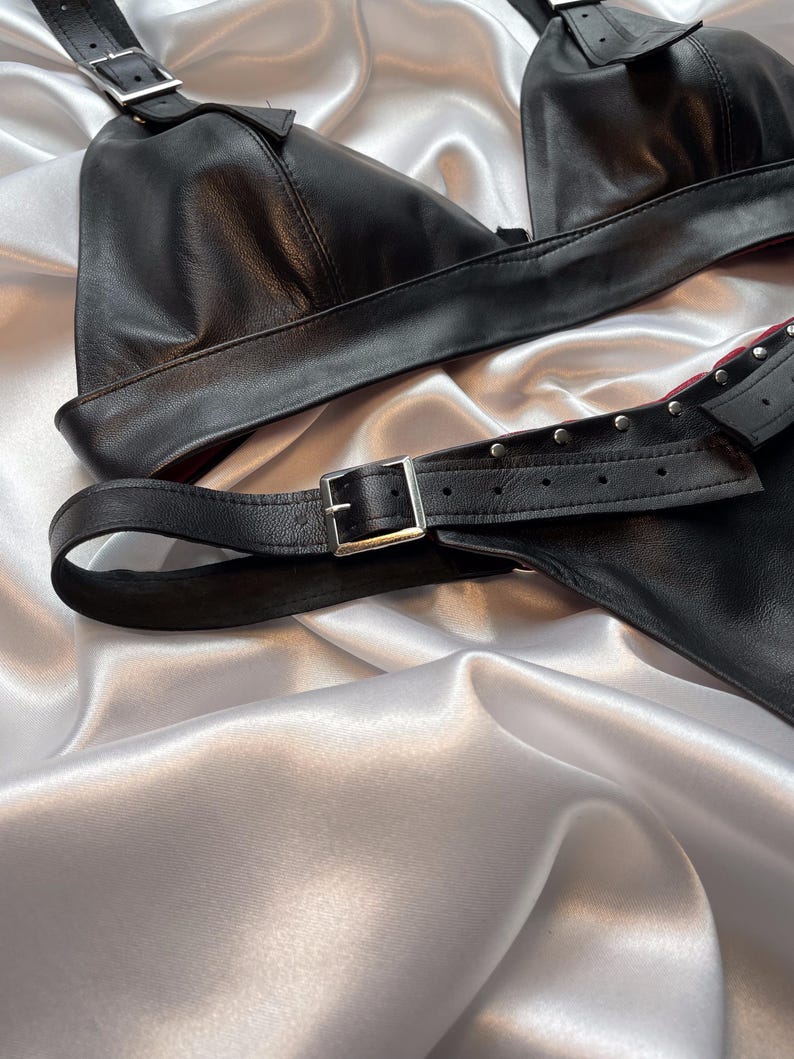 May include: Black leather lingerie set with adjustable straps and silver-tone buckles. The set includes a triangle-shaped top and a matching bottom piece. The lingerie is displayed on a white satin surface.
