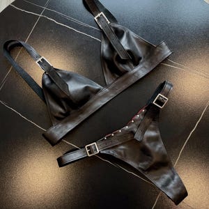 May include: Black leather lingerie set featuring a triangle-shaped bra and a thong. Both pieces have adjustable straps with silver-colored buckles. The thong has a red interior and is embellished with small silver studs.