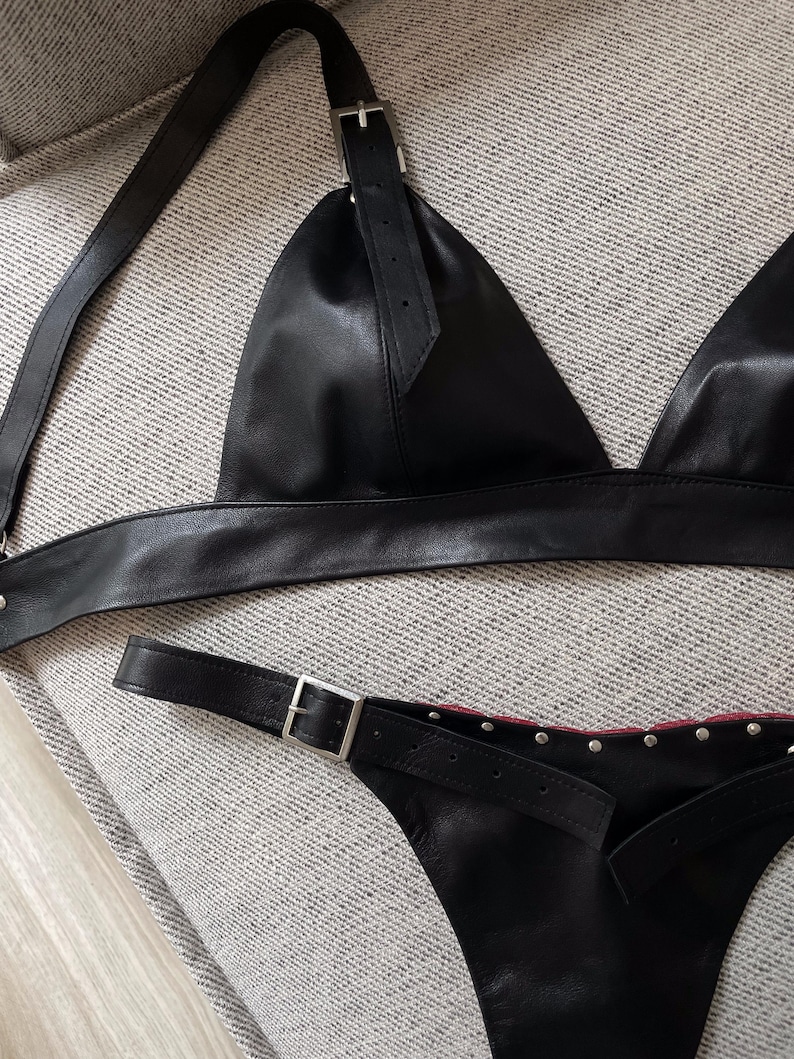 May include: Black leather lingerie set featuring a triangle top with adjustable straps and a matching bottom. The set includes silver-toned metal buckles and rivets. The lingerie is displayed on a neutral-toned surface.