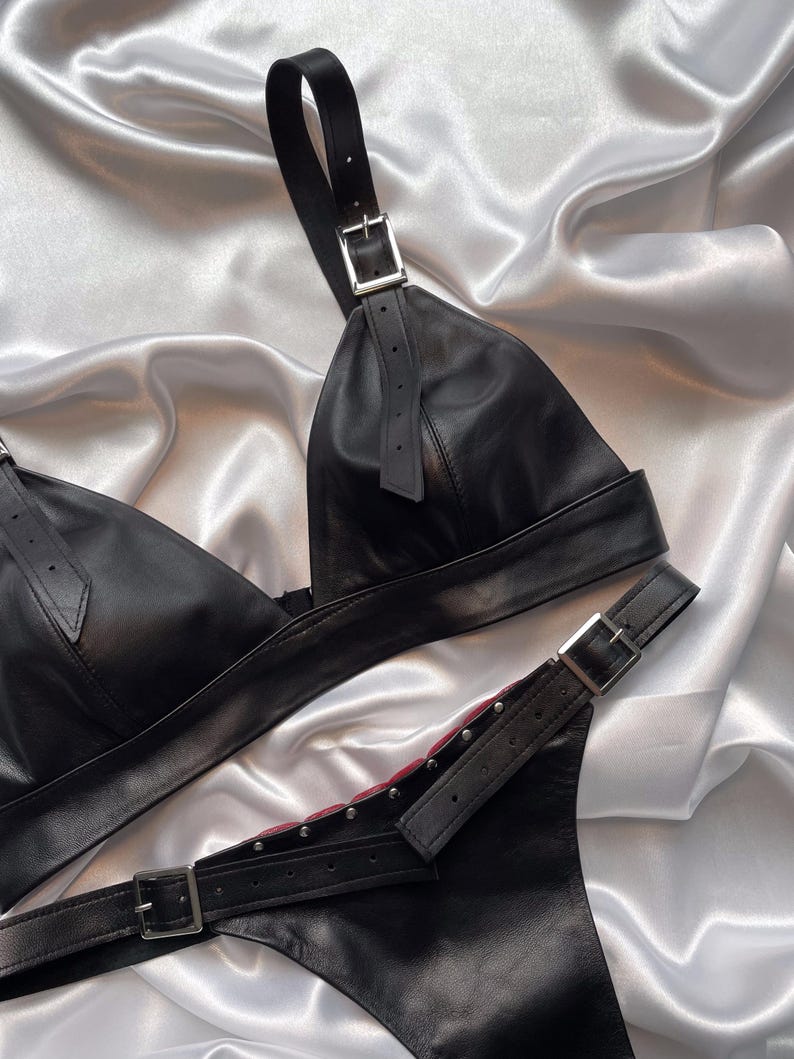 May include: Black leather lingerie set featuring a triangle bra with adjustable straps and silver buckles. The matching bottom has a buckle closure and silver stud details. The set is displayed on a white and silver satin background.