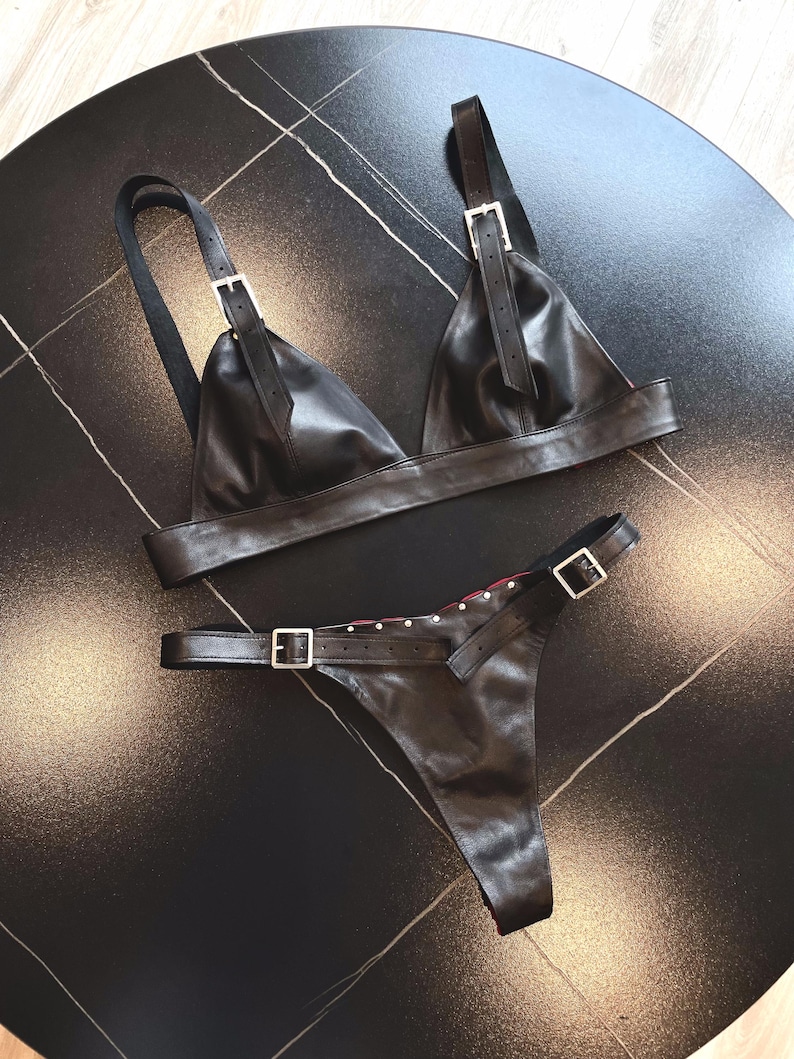 May include: Black leather lingerie set. The set includes a triangle-style bra with adjustable straps and a matching thong. Both pieces feature silver-tone metal buckles and hardware. The thong has decorative studs along the waistband.