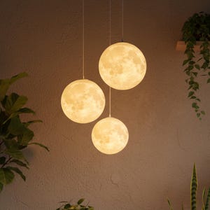 Könnte beinhalten: Drei kugelförmige Pendelleuchten mit Mondoberfläche hängen von der Decke. Die Lichter sind beleuchtet und erzeugen ein warmes Leuchten. Die Szene umfasst Pflanzen und einen Holztisch und schafft eine gemütliche Atmosphäre.