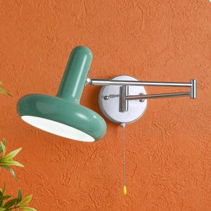 Könnte beinhalten: Eine grüne Wandleuchte mit weißem Innenraum und Zugkette. Die Lampe ist auf einer weißen, kreisförmigen Basis mit einem silbernen, verstellbaren Arm montiert. Der Hintergrund ist eine orangefarbene, strukturierte Wand.