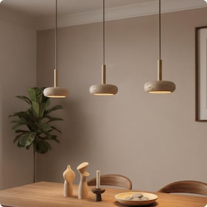 Peut inclure: Trois lampes suspendues modernes avec un abat-jour circulaire beige et texturé et une tige cylindrique en bois. Les lampes sont suspendues au-dessus d'une table à manger en bois avec des objets décoratifs en bois et une bougie. La pièce a une palette de couleurs neutres.